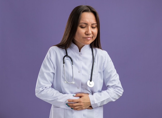 Foto gratuita giovane dottoressa asiatica dolorante che indossa una tunica medica e uno stetoscopio che guarda in basso tenendo le mani sul ventre isolato sul muro viola con spazio di copia