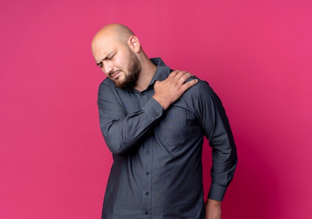 Aching young bald call center man putting hand on shoulder with closed eyes isolated on crimson  with copy space