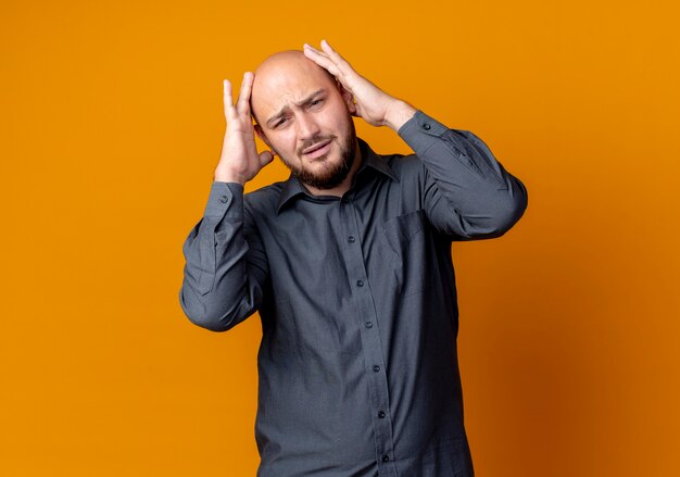 Aching young bald call center man putting hands on head looking straight isolated on orange  with copy space