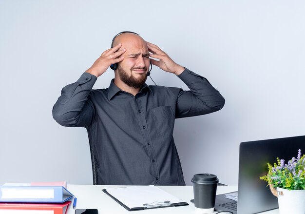 Aching young bald call center man wearing headset sitting at desk with work tools looking at side putting hands on head isolated on white 