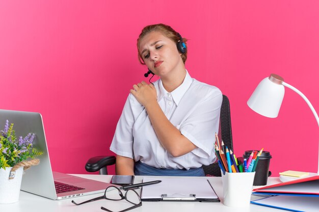 Aching young blonde call centre girl wearing headset sitting at desk with work tools keeping hand on shoulder looking at laptop isolated on pink wall