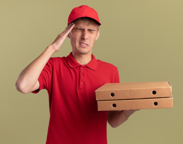 Aching young blonde delivery boy puts hand on forehead and holds pizza boxes isolated on olive green wall with copy space