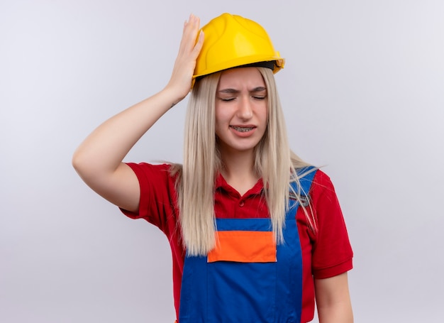 Бесплатное фото Болит молодая блондинка инженер-строитель девушка в униформе в зубных скобах, положив руку на голову с закрытыми глазами на изолированном белом пространстве