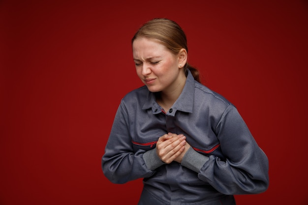 目を閉じて胸に手を保つ制服を着て痛む若いブロンドの女性エンジニア