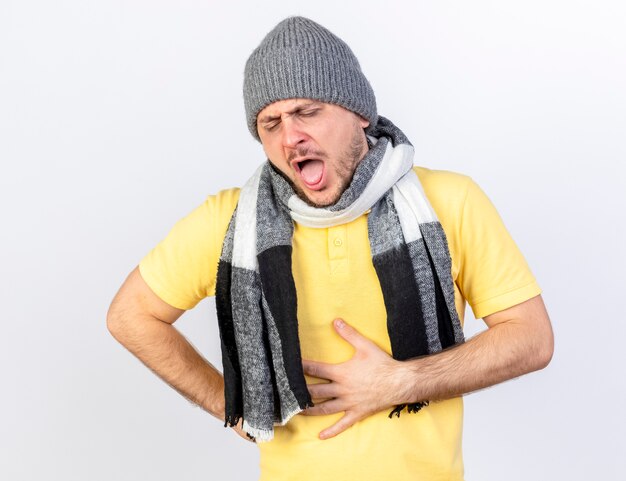 Aching young blonde ill man wearing winter hat and scarf puts hand on belly isolated on white wall