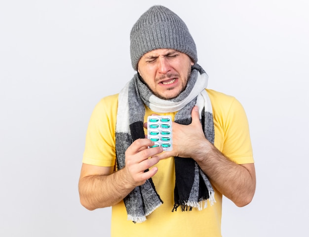 Aching young blonde ill man wearing winter hat and scarf puts hand on chest and holds pack of medical pills isolated on white wall