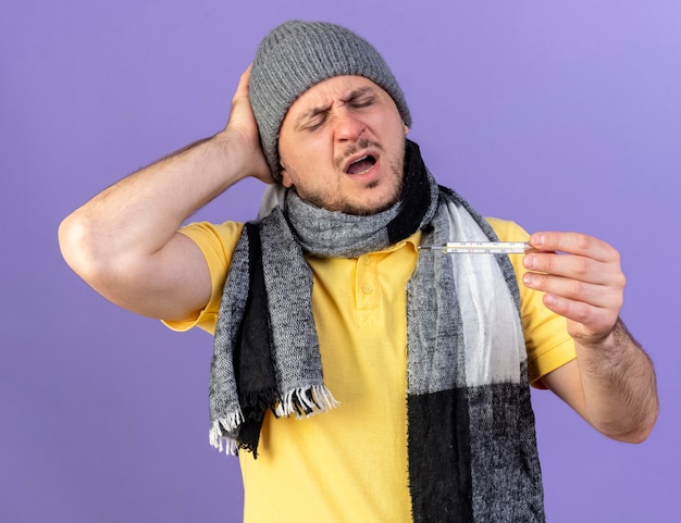 Aching young blonde ill man wearing winter hat and scarf puts hand on head behind looking at thermometer isolated on purple wall