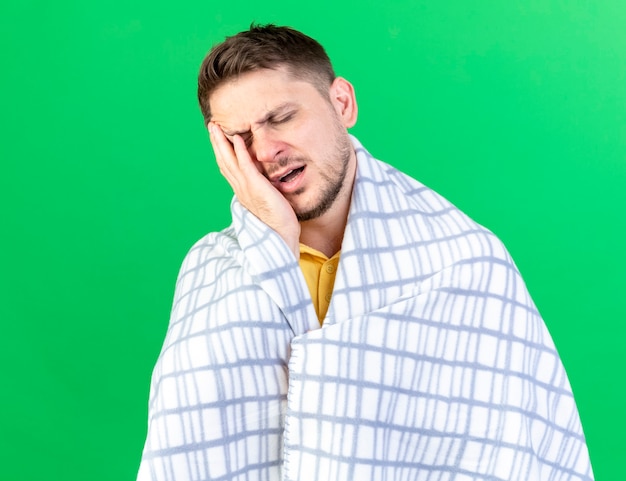 Free photo aching young blonde ill man wrapped in plaid puts hand on face isolated on green wall