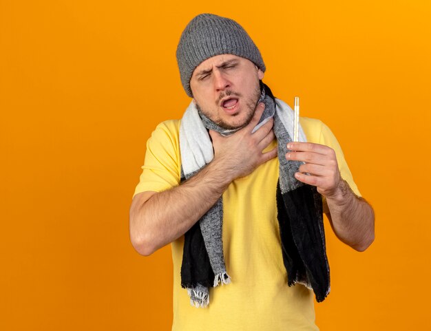 Aching young blonde ill slavic man wearing winter hat and scarf puts hand on neck