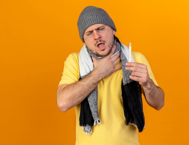 Больной молодой блондин больной славянский мужчина в зимней шапке и шарфе кладет руку на шею