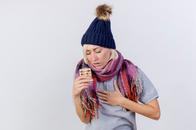 Aching young blonde ill slavic woman wearing winter hat and scarf puts hand on chest and holds pack of medical pills isolated on white wall with copy space