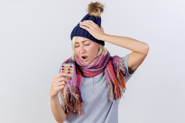 冬の帽子とスカーフを身に着けている痛む若い金髪の病気のスラブ女性は頭に手を置き、コピースペースで白い壁に分離された医療薬のパックを保持します