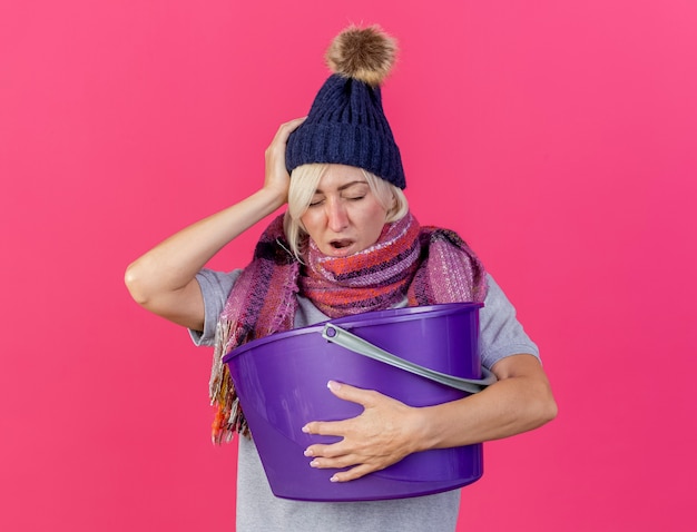 Aching young blonde ill slavic woman wearing winter hat and scarf puts hand on head holds plastic bucket isolated on pink wall with copy space