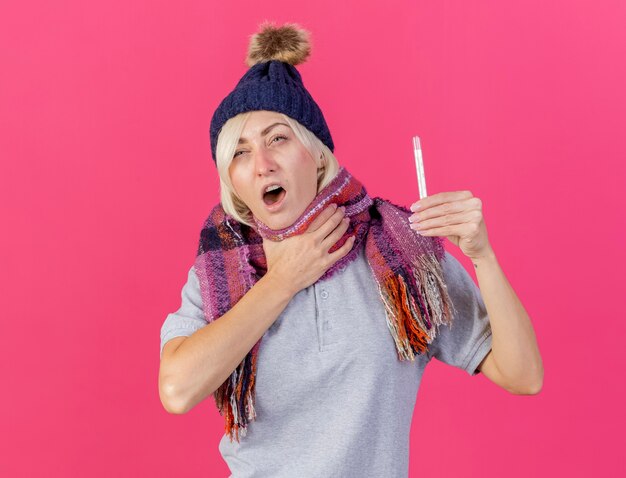 冬の帽子とスカーフを身に着けている痛む若い金髪の病気のスラブ女性は首に手を置き、コピースペースでピンクの壁に隔離された温度計を保持します