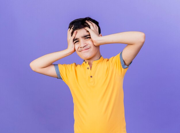 Aching young boy looking at front holding head isolated on purple wall