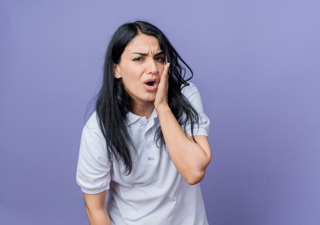 Aching young brunette caucasian girl puts hand on face isolated on purple wall