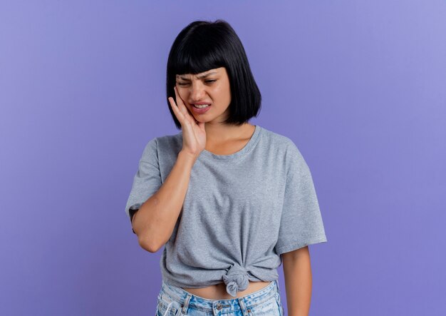 Aching young brunette caucasian woman puts hand on face isolated on purple background with copy space