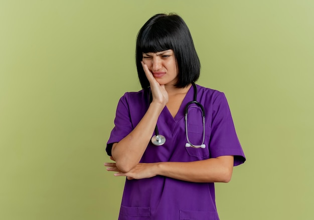 Foto gratuita dolorante giovane donna bruna medico in uniforme con lo stetoscopio mette la mano sul viso isolato su sfondo verde oliva con lo spazio della copia