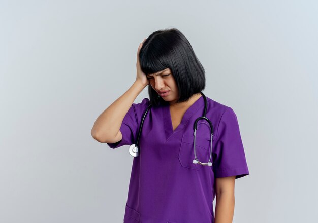 Aching young brunette female doctor in uniform with stethoscope puts hand on head