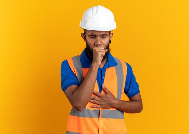 aching young builder man in uniform with safety helmet coughing isolated on orange wall with copy space