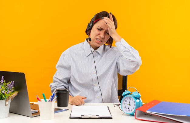 Aching young call center girl wearing headset sitting at desk putting hand on head with closed eyes isolated on orange 