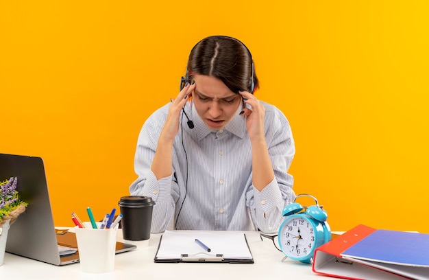 Dolorante giovane ragazza della call center che indossa la cuffia avricolare che si siede allo scrittorio che mette le mani sulla testa che soffrono di mal di testa con gli occhi chiusi isolati sull'arancio