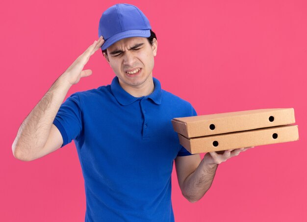 Болит молодой кавказский курьер в синей форме и кепке, держащий упаковки с пиццей, касаясь головы, страдающий от головной боли с закрытыми глазами