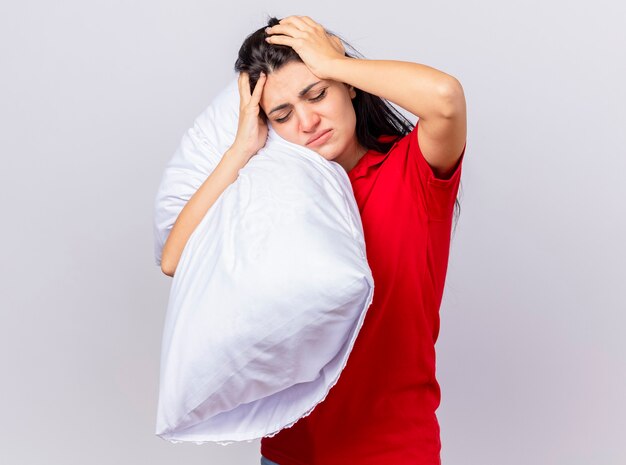 Aching young caucasian ill girl holding pillow and head with closed eyes isolated on white background with copy space