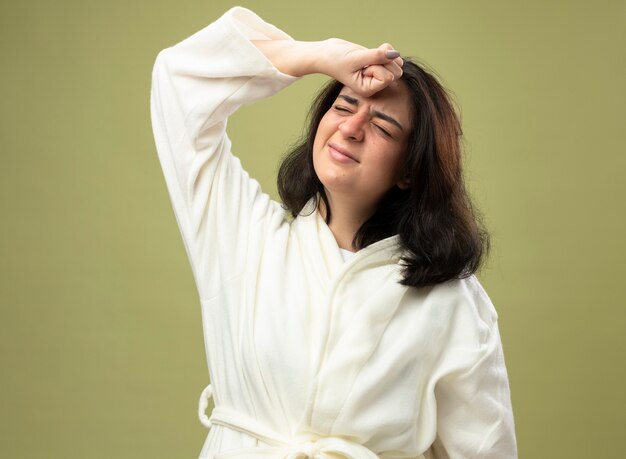 Aching young caucasian ill girl wearing robe keeping fist on forehead with closed eyes isolated on olive green background with copy space