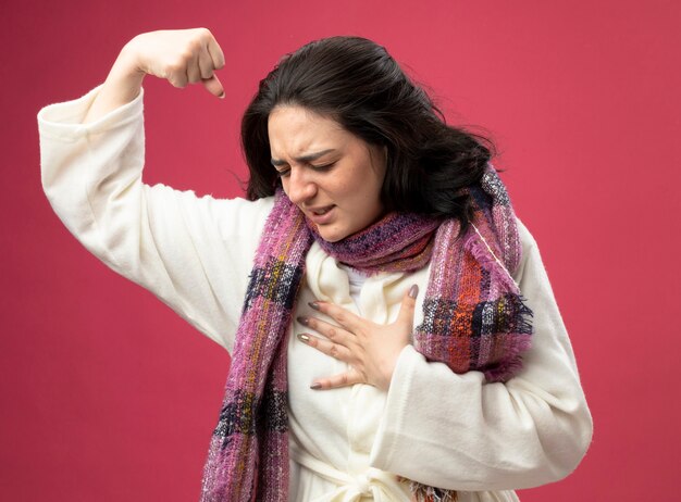 Aching young caucasian ill girl wearing robe and scarf putting hand on chest doing strong gesture with closed eyes isolated on crimson wall