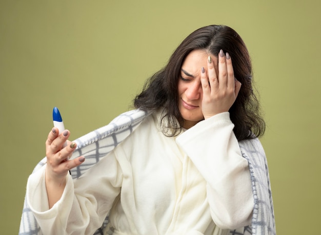 Aching young caucasian ill girl wearing robe wrapped in plaid holding thermometer putting hand on head with closed eyes isolated on olive green background