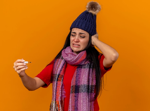 Aching young caucasian ill girl wearing winter hat and scarf holding and looking at thermometer putting hand on head isolated on orange wall with copy space