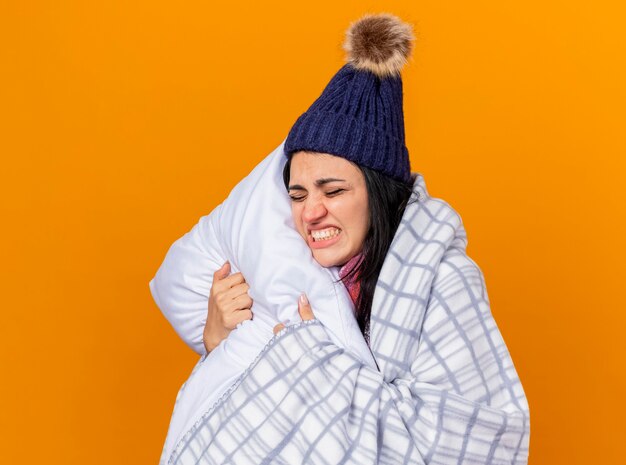 Aching young caucasian ill girl wearing winter hat and scarf wrapped in plaid hugging pillow with closed eyes isolated on orange background with copy space