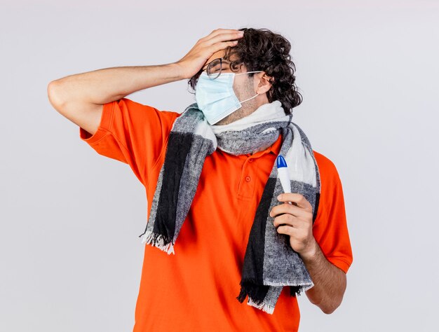 Aching young caucasian ill man wearing glasses mask and scarf holding thermometer putting hand on head with closed eyes isolated on white wall