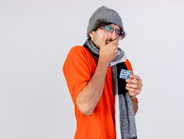 Aching young caucasian ill man wearing glasses winter hat and scarf holding pack of medical capsules keeping hand on mouth having toothache isolated on white background with copy space