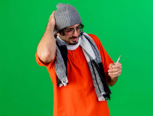 Aching young caucasian ill man wearing glasses winter hat and scarf holding thermometer keeping hand on head isolated on green background with copy space