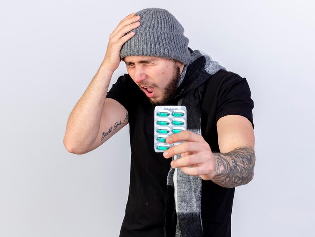 Aching young caucasian ill man wearing winter hat and scarf puts hand on head