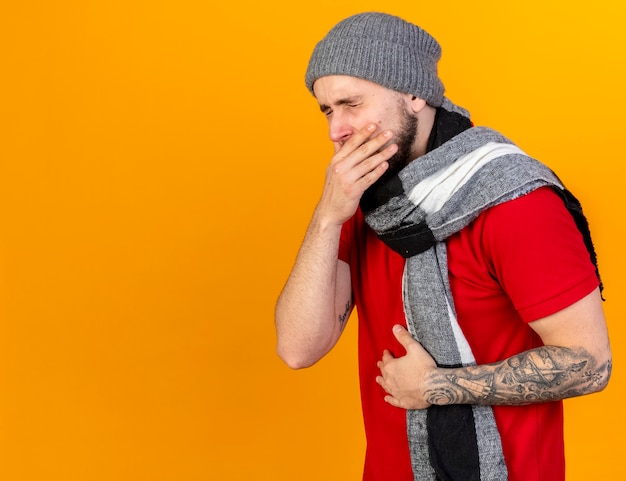 Aching young caucasian ill man wearing winter hat and scarf puts hand on mouth and belly isolated on orange wall with copy space