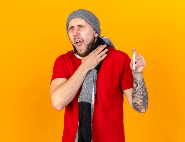Aching young caucasian ill man wearing winter hat and scarf puts hand on neck and holds thermometer