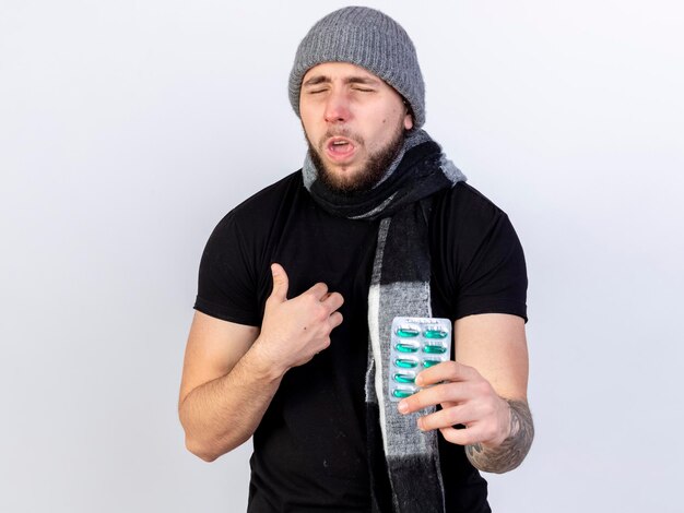 Aching young caucasian ill man wearing winter hat and scarf stands with closed eyes