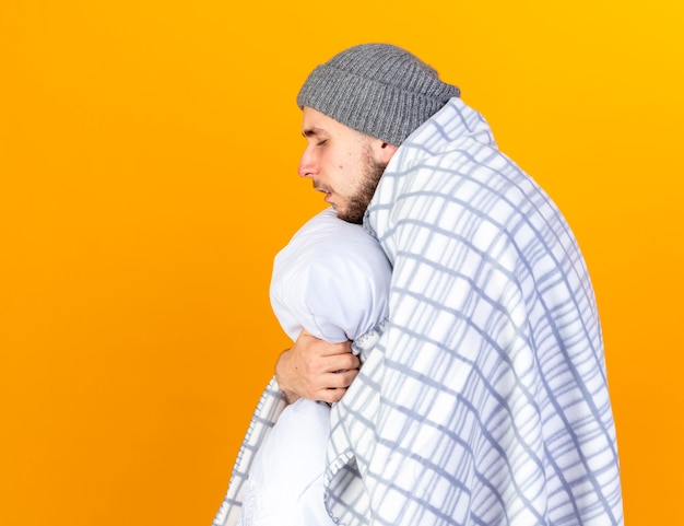 Aching young caucasian ill man wearing winter hat and scarf wrapped in plaid stands sideways holding pillow isolated on orange wall with copy space