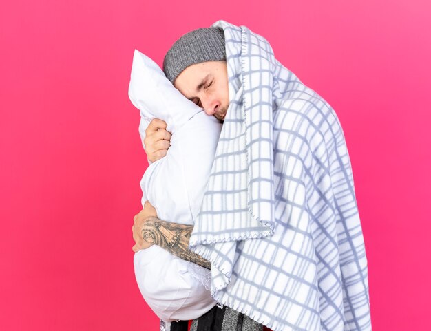 Aching young caucasian ill man wearing winter hat wrapped in plaid hugs and puts head on pillow isolated on pink wall with copy space