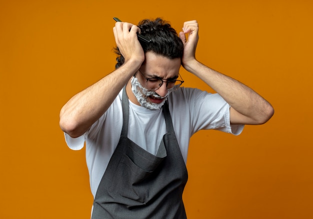 Giovane barbiere maschio caucasico dolorante che indossa occhiali e fascia per capelli ondulati in uniforme che tiene un rasoio con crema da barba messo sul viso tenendo la testa che soffre di mal di testa con gli occhi chiusi