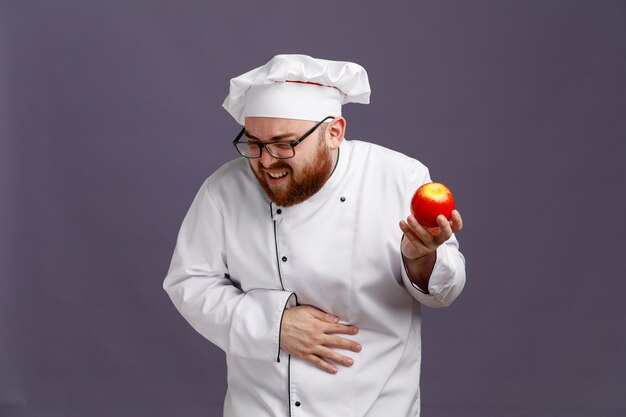 Больной молодой шеф-повар в очках и кепке держит яблоко, держа руку на животе, глядя вниз на фиолетовом фоне