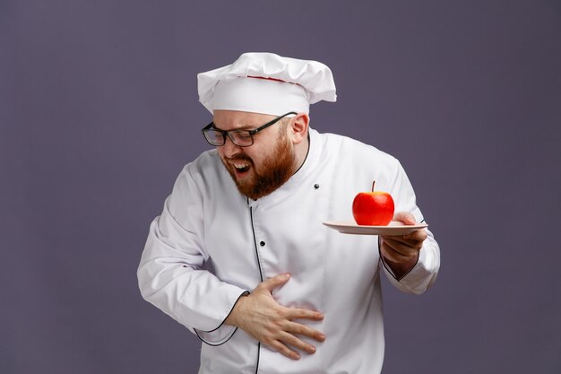 aching young chef wearing glasses uniform and cap holding apple in plate keeping hand on belly with closed eyes isolated on purple background
