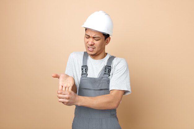 Aching young construction worker wearing safety helmet and uniform holding hand with closed eyes 