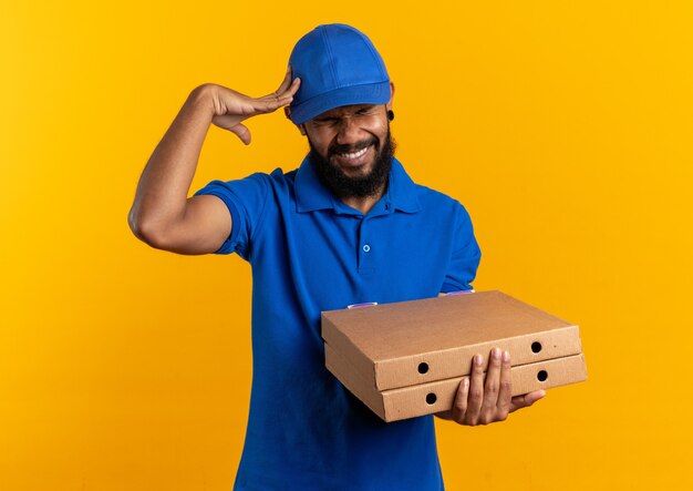 aching young delivery man holding pizza boxes and putting hand on his head isolated on orange wall with copy space