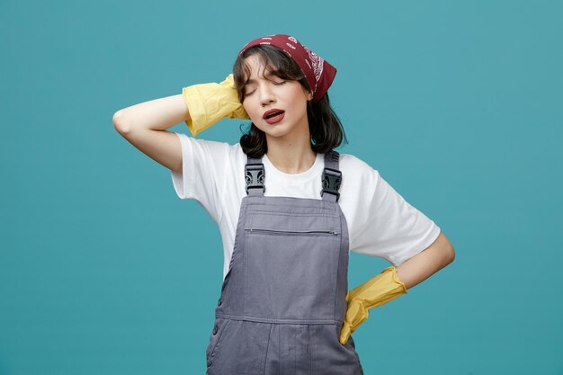 Aching young female cleaner wearing uniform bandana and rubber gloves keeping hand on head and on waist with closed eyes isolated on blue background