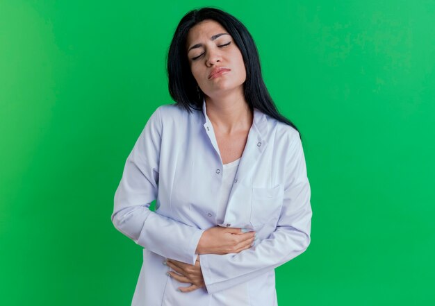 コピースペースと緑の壁に隔離された目を閉じて腹を保持している医療ローブを身に着けている痛む若い女性医師