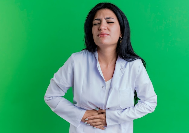 Aching young female doctor wearing medical robe holding belly with closed eyes 